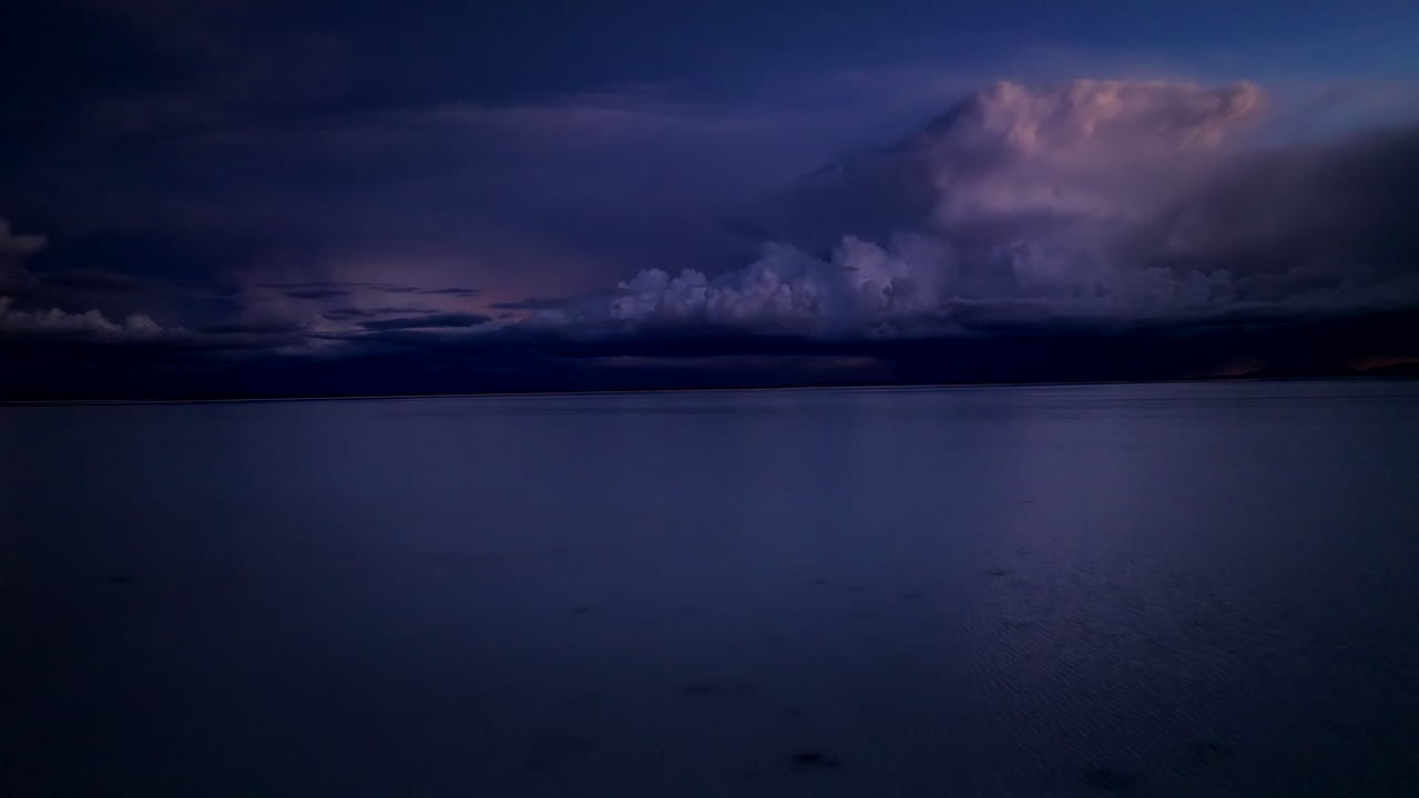 Aerial view over Uyuni salt flats at twilight. Deep blue tones and low cloud formations create a surreal and calm mirrored scene on the water’s surface