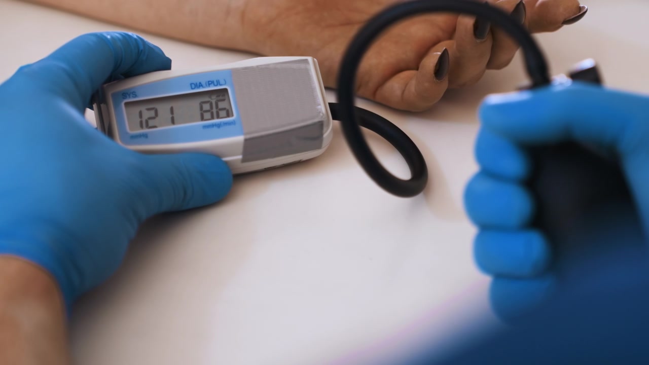 Controllo della pressione sanguigna a domicilio di un paziente di colore da parte di un infermiere paramedico che indossa guanti in nitrile blu, usando un bracciale digitale e una pompetta di gonfiaggio collegati a un monitor al polso, scena tranquilla in una residenza interna con un infermiere attento.