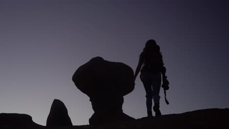 Silhouette-of-Female-Hiker-With-Backpack-and-Photo-Camera-Hiking-Up-on-Hill-With-Strange-Rock-Formation,-Slow-Motion-Full-Frame