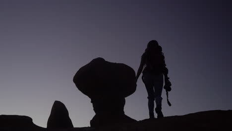 silhouette of female hiker with backpack and photo camera hiking up on hill with strange rock formation, slow motion full frame