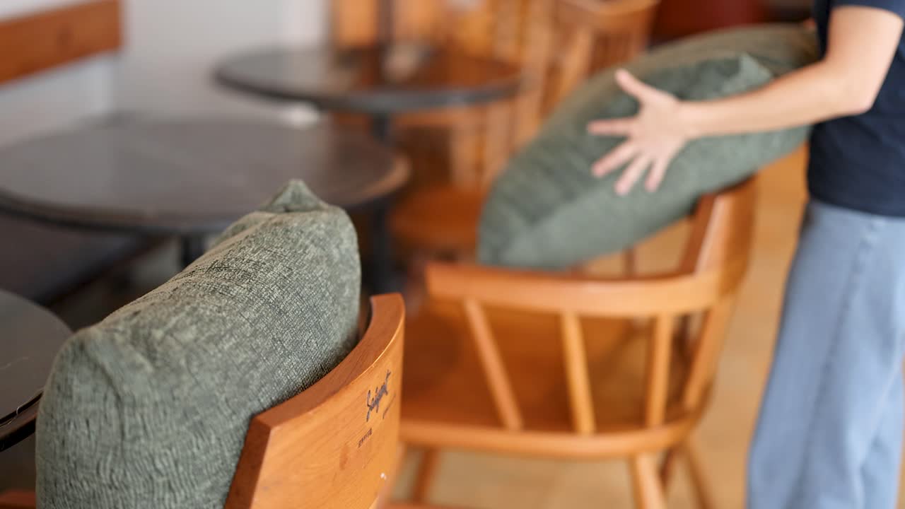 Person arranges green cushion on wooden chair in softly lit, modern cafe with wooden furniture