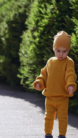 lovely kid wearing yellow knitted suit stands outdoors. baby boy looks at something down with interest. vertical video