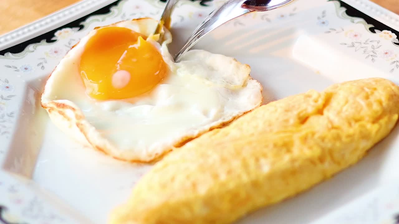Close-up of a fork slicing into a fried egg yolk beside a golden omelet on a decorative plate.