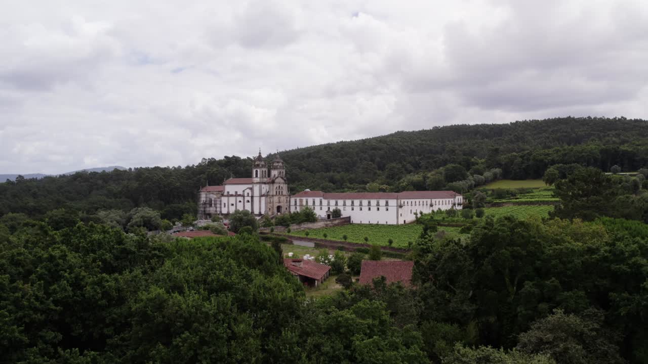 Vista aérea do Mosteiro de São Martinho de Tibães com vinhedos, campos e colinas arborizadas circundantes sob céu nublado.