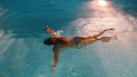 Slow-motion:-Young-woman-swimming-by-herself-inside-of-a-pool-at-night-time,-pool-lights-are-lighting-up-the-sides-of-the-pool