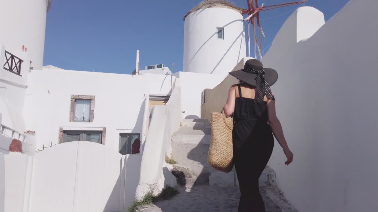 joven con un elegante traje negro y sombrero, caminando en la ciudad de oia, isla de santorini, grecia, cámara lenta