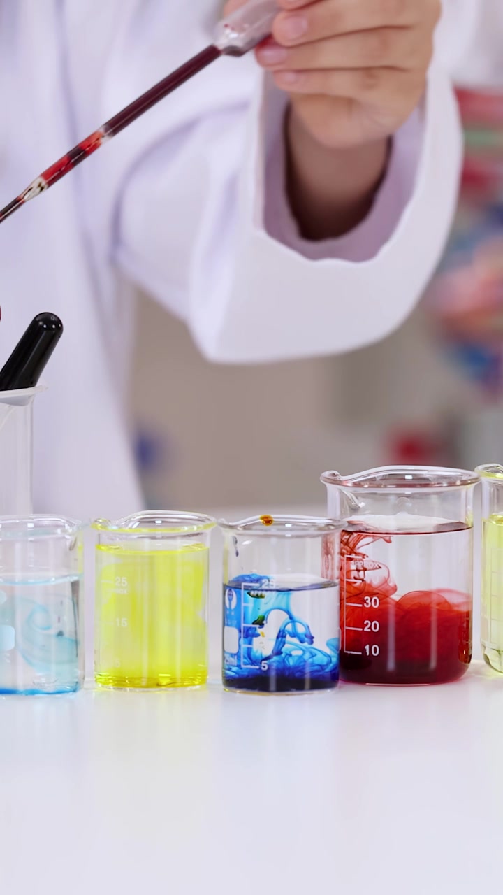 Primo piano di uno studente in camice bianco che aggiunge liquido colorato a becher di vetro.