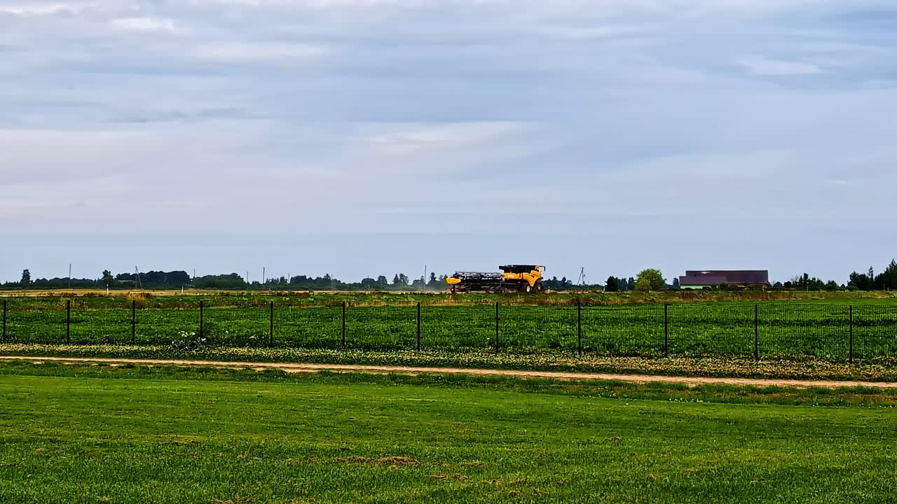Yellow harvester operates across open green fields under cloudy skies near Dobele