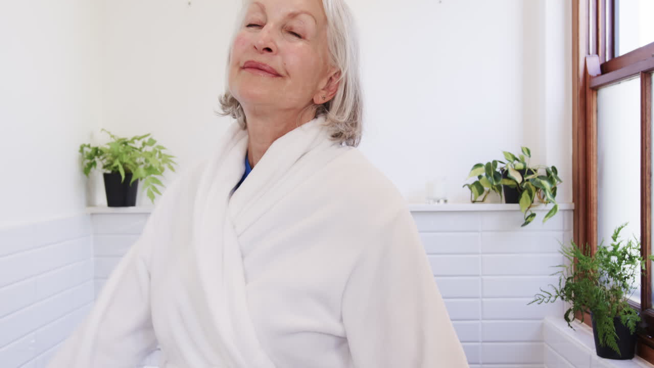 Senior woman enjoying peaceful moment in bathroom, wearing white robe, smiling, at home