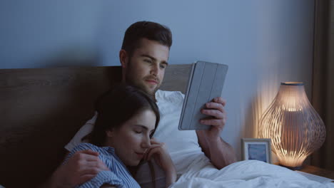 young caucasian couple sitting the bed and watching a video on the tablet device at night