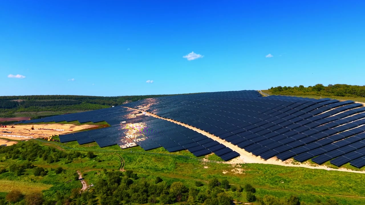 Solcellepark under opførelse på en bjergskråning omgivet af grøn vegetation. Droneudsigt over vinklede paneler med solrefleksion, skov og byggeudstyr på en klar dag.