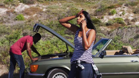 african american woman talking on smartphone while her husband trying to fix the car on road