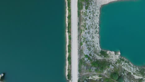 Aerial-top-down-narrow-bridge-road-over-natural-lake-between-mountain-pine-tree-forest