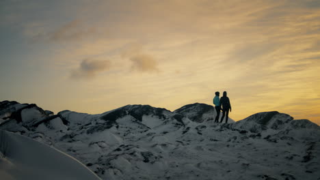 Panning-shot-of-a-couple-standing-at-the-top-of-a-snowy-Lovstakken-at-sunrise