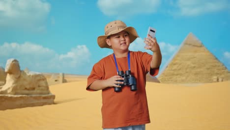 asian boy with a hat and binoculars waving hand having video call on smartphone while traveling in giza pyramid. boy researcher examines something, travel tourism adventure