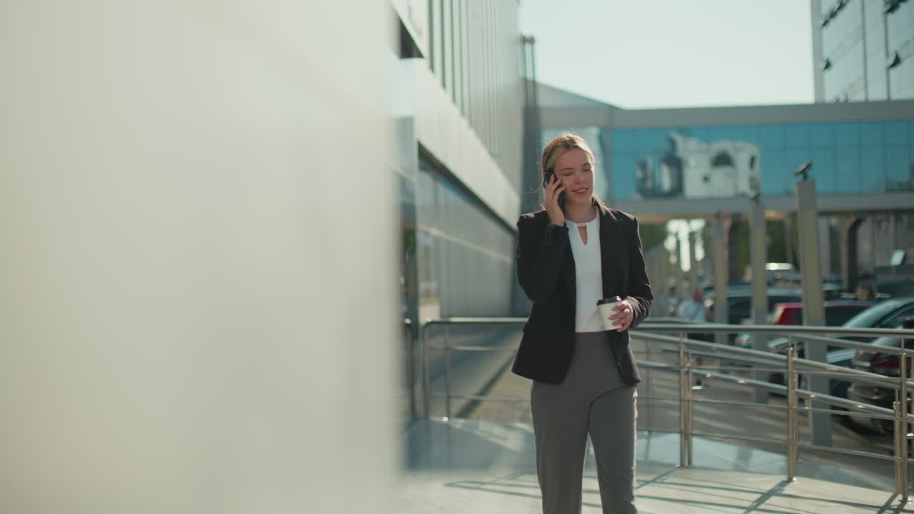 Lecturer walking confidently with coffee in hand on phone call near iron railing, with blur background featuring parked cars, glass building reflections, and person standing under sunlight
