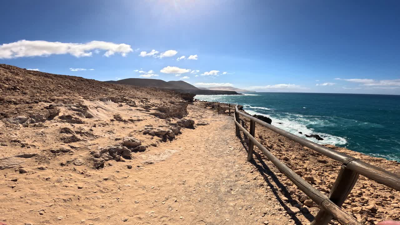 Visuale soggettiva a piedi lungo le aspre scogliere vulcaniche e il terreno costiero roccioso di Fuerteventura, Spagna, sotto un cielo sereno e soleggiato in estate.