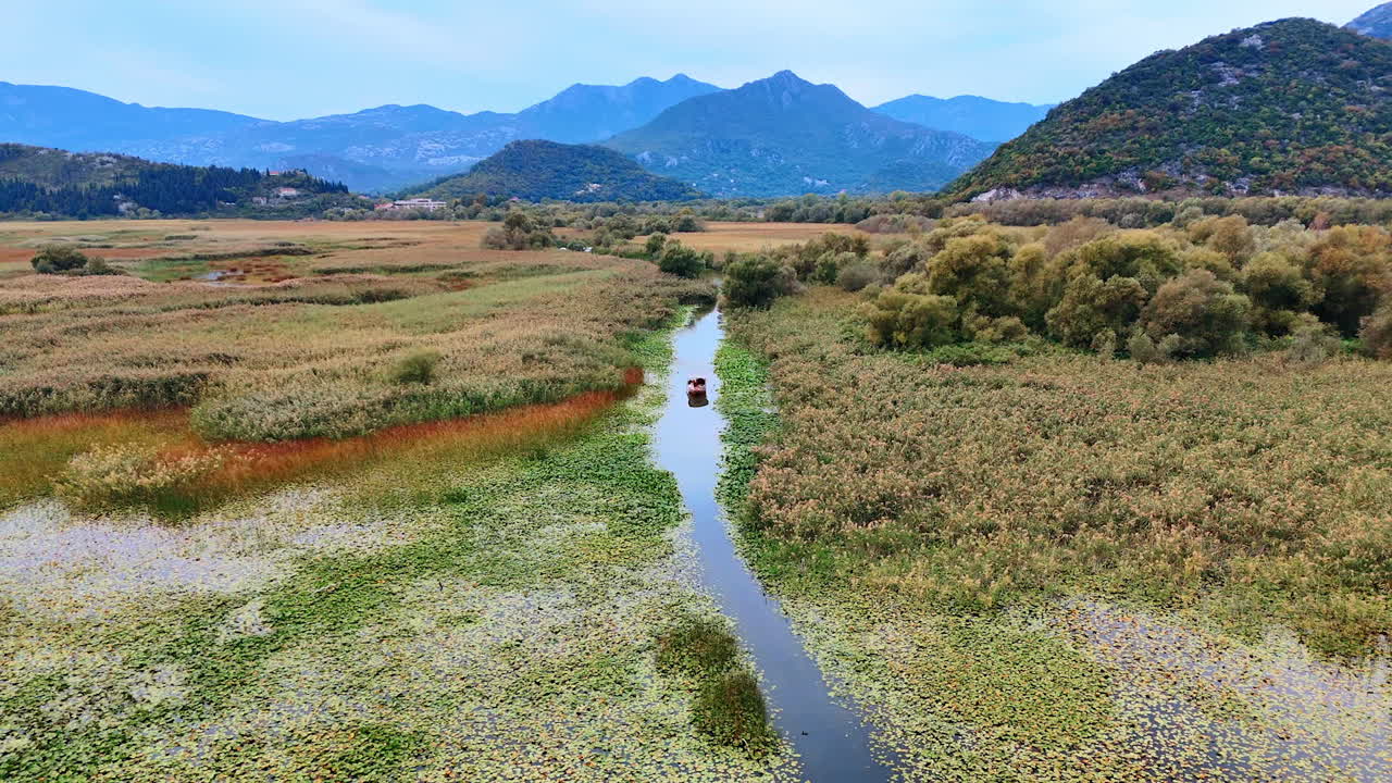 Boat moves by the narrow river overgrown with weeds. Beautiful nature landscapes and mountains range at backdrop