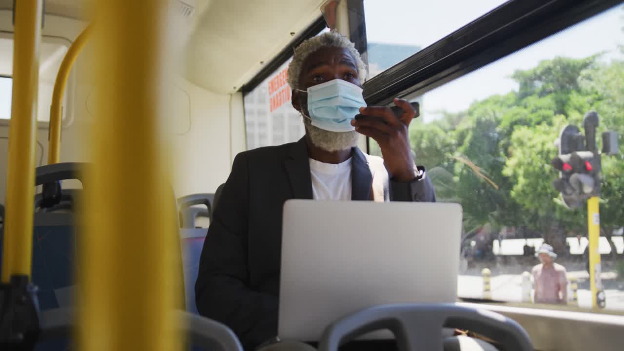 Afroamerikansk ældre mand iført mundbind taler i smartphone og bruger bærbar computer, mens han sidder.