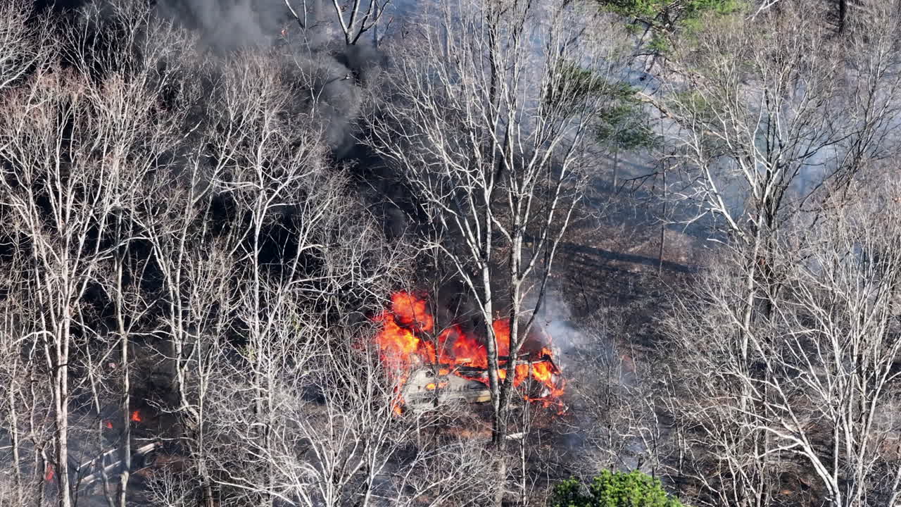 Telephoto aerial drone footage of a camper van that is on fire in the forest.