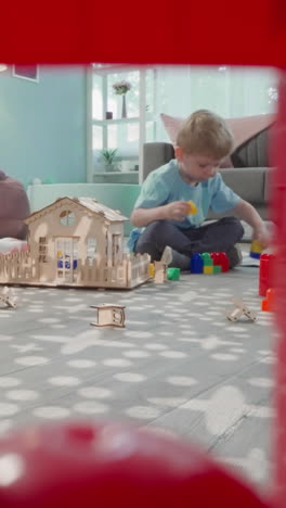 cute toddler plays with plastic blocks after building wooden house. blond boy develops new skills at home view through clearance in construction of cubes