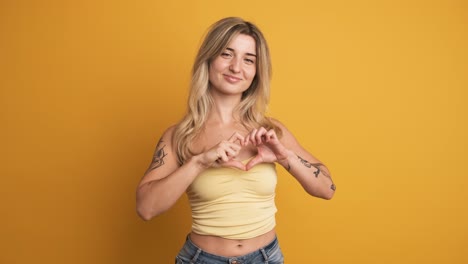 happy young woman sending flying kiss and showing heart sign with hand on yellow background