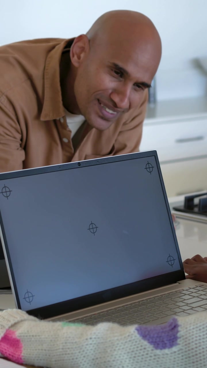 Vertikales Video: Mann im braunen Hemd lehnt sich, zeigt auf einen markierten Laptop in der Küche und präsentiert einen cremefarbenen Pullover.