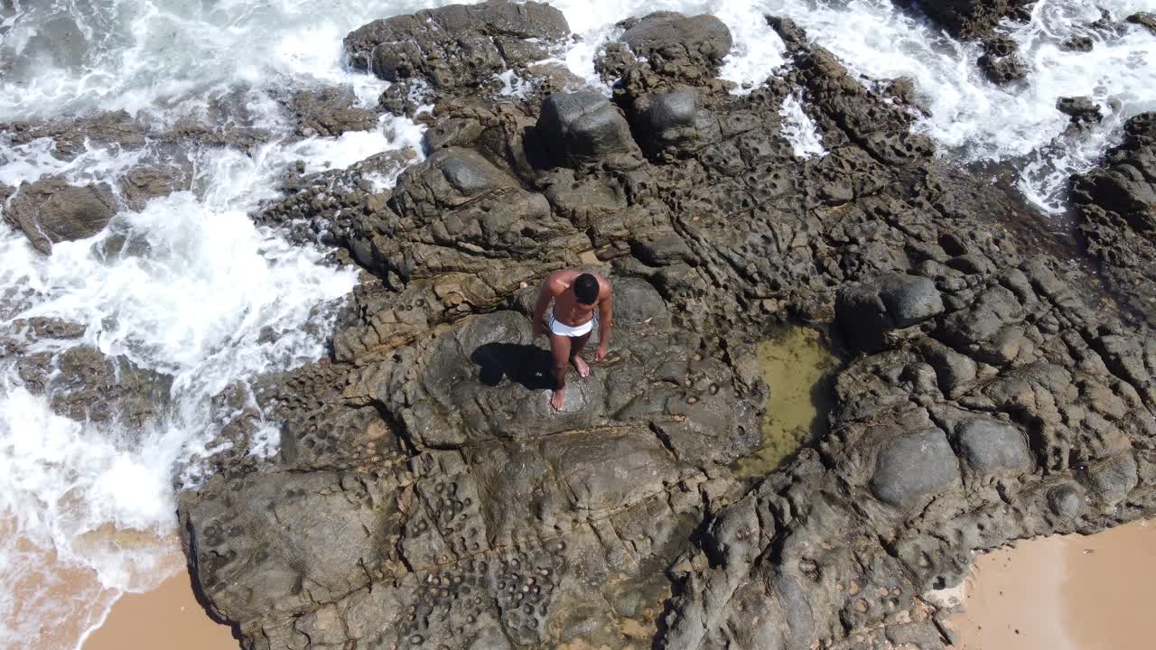 A drone ascends, revealing a Black man on a rock and a vast beach coastline.