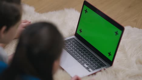 chromakey laptop screen with relaxed teenage boy and girl watching. green screen device on floor at home indoors with caucasian friends siblings enjoying leisure. mockup template.
