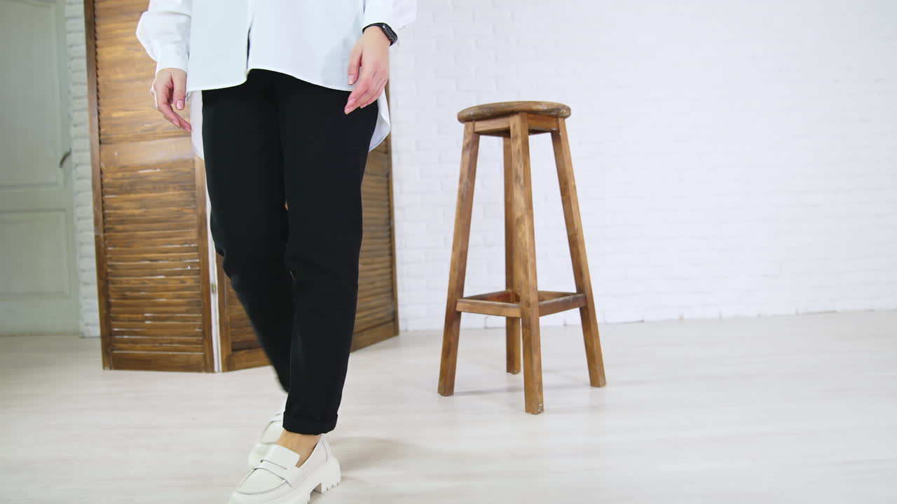 Lady in white shirt and black jeans demonstrates white fashionable shoes. Woman walks in plain loafers with massive soles back and forth near wooden tabouret.