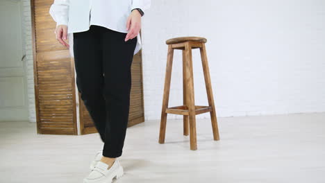 lady in white shirt and black jeans demonstrates white fashionable shoes. woman walks in plain loafers with massive soles back and forth near wooden tabouret.