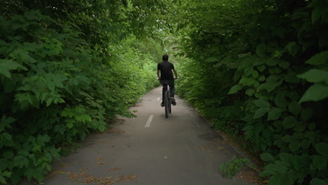 back view of young boy in black top and grey trouser riding his bike along a paved path with white markings, surrounded by dense greenery with leaves littered on the walk path