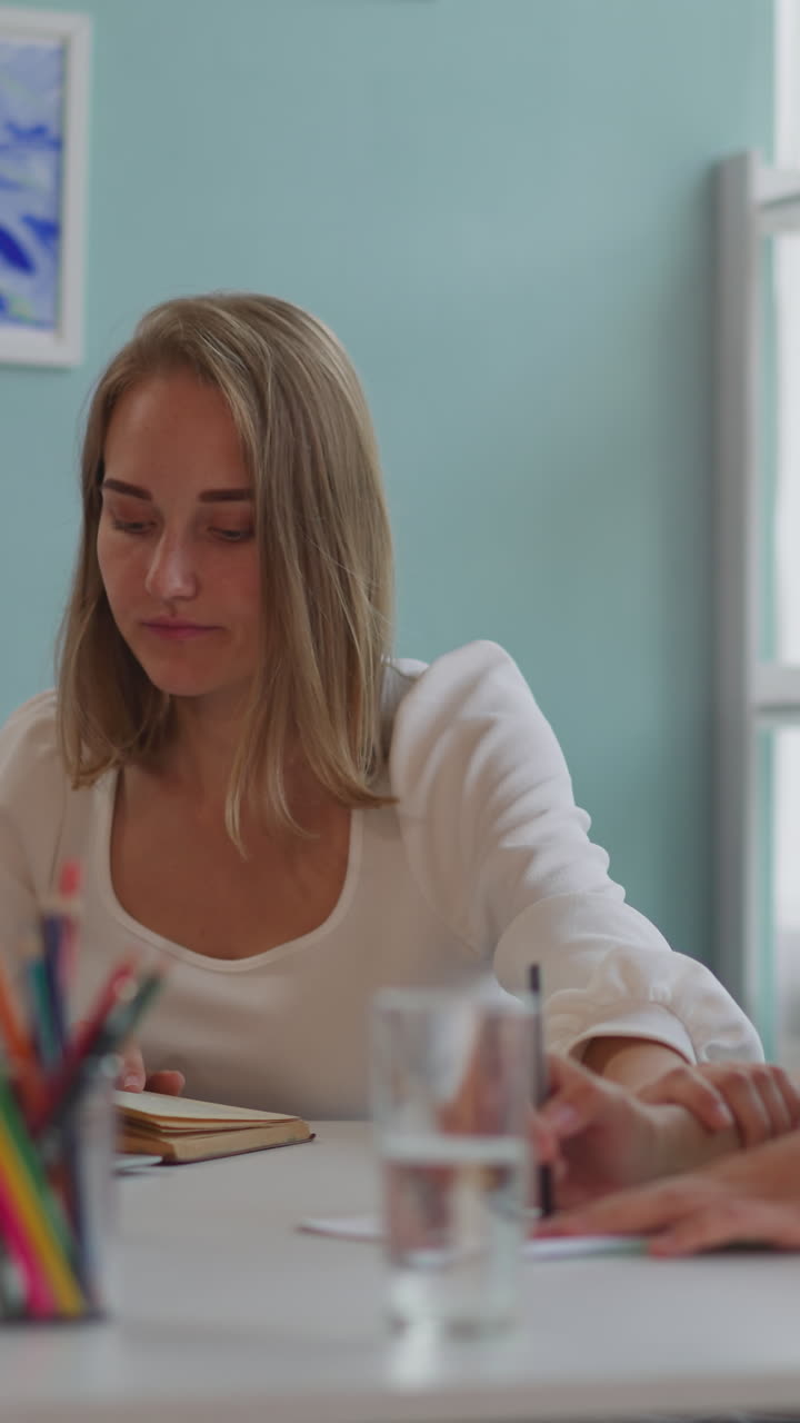 Young woman touches hand of girl partner with disability to express love while doing homework. Ladies prepare for classes sitting at table closeup