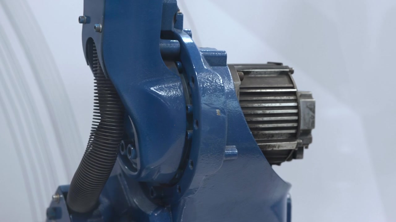 Blue industrial machine assembly shown in close side view with glossy painted housing, ribbed motor section, corrugated cable, bolts, and precise joints against soft pale background, creating clean
