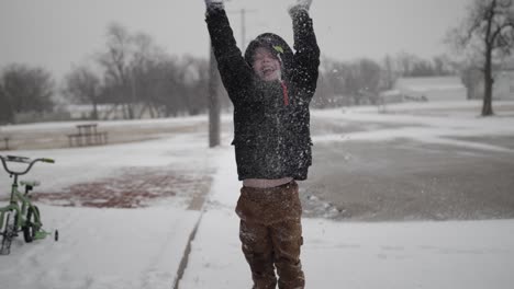 joven, feliz, niño sonriendo y usando un abrigo jugando afuera en un frío día de invierno en diciembre arroja nieve al aire durante las vacaciones de navidad en un pequeño pueblo en el medio oeste