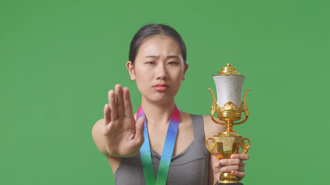 금메달과 트로피를 들고 있는 아시아 여성의 클로즈업, 손짓 없이 불쾌감을 표하며 스튜디오의 녹색 스크린 배경에서 카메라에 미소 짓고 있다.