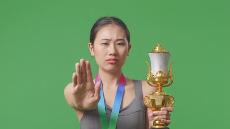 primer plano de una mujer asiática con una medalla de oro y un trofeo desaprobando sin signos de mano y sonriendo a la cámara en el fondo de la pantalla verde en el estudio