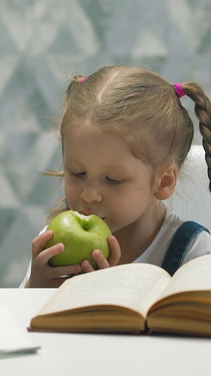 slow motion portrait of little girl during self-isolation and home schooling takes break, child eats delicious green apple, sits at table with book notebook and scissors