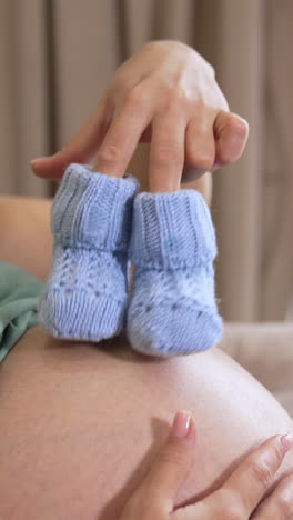 pregnant woman playing with tiny baby socks. future mom with blue knitted socks stepping on her belly while relaxing at home. expectant mother enjoys pregnancy time. vertical video