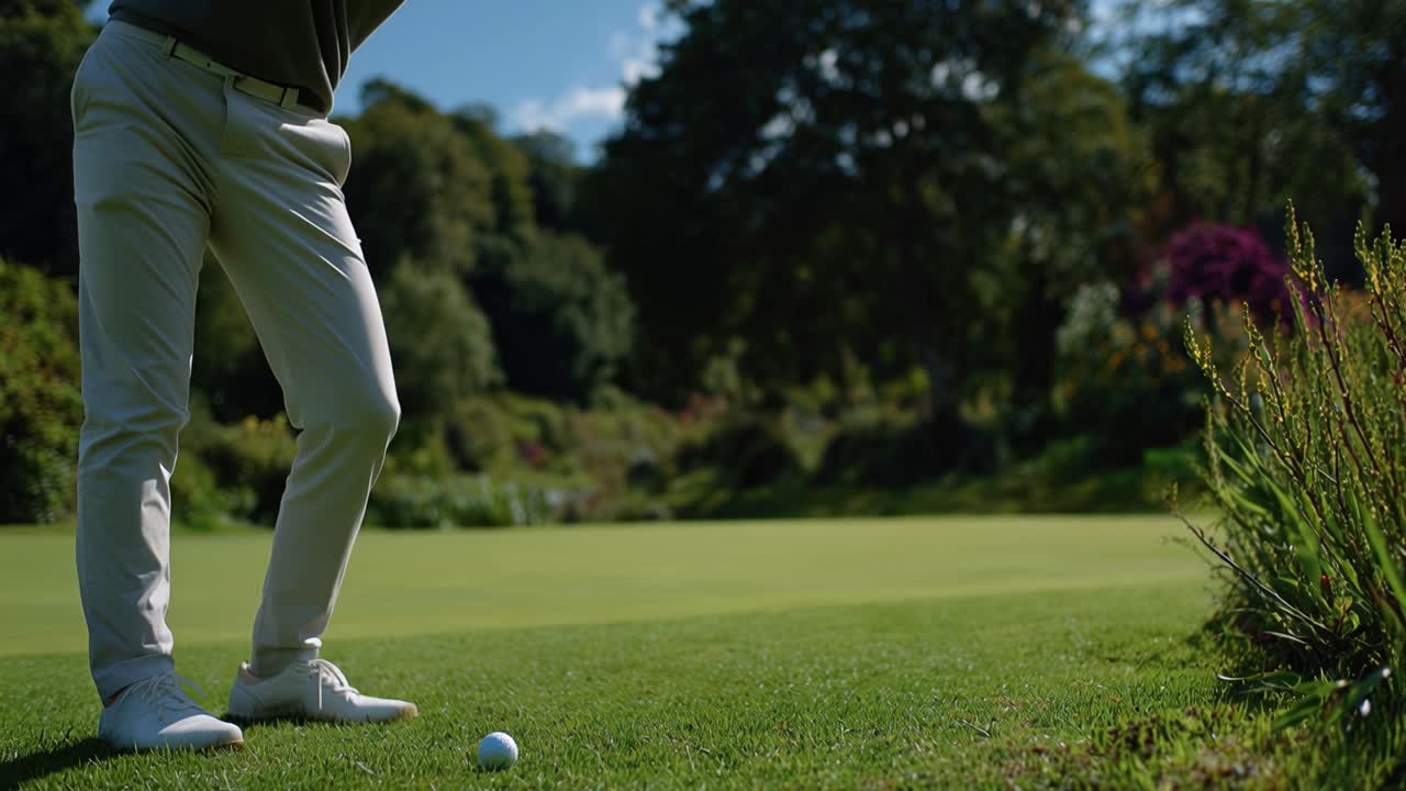 Golfista se připravuje k patování na greenu. Jsou vidět bílé kalhoty, golfové boty a hůl. Sluneční světlo vytváří jasné stíny na trávě. Hráč se soustředí na míček a má stabilní postoj.