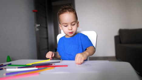 calm little kid sitting at desk busy with drawing. toddler pulls the sheet of paper closer holding a brown pencil.