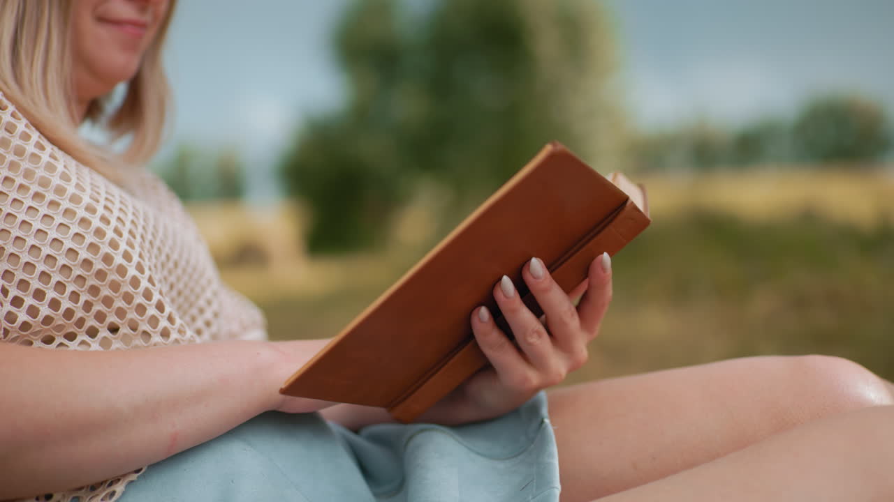 primer plano de una mujer leyendo un libro viejo al aire libre, volviendo delicadamente las páginas con manos bien cuidadas, enfoque suave en pantalones cortos azules, fondo natural borroso
