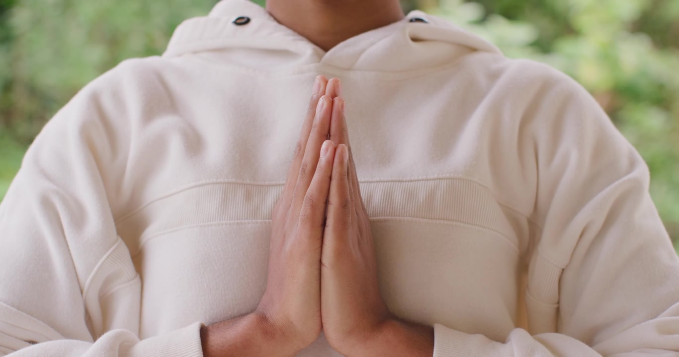 African woman starting sequence holding palms pressed at chest, meditating in garden wearing hoodie