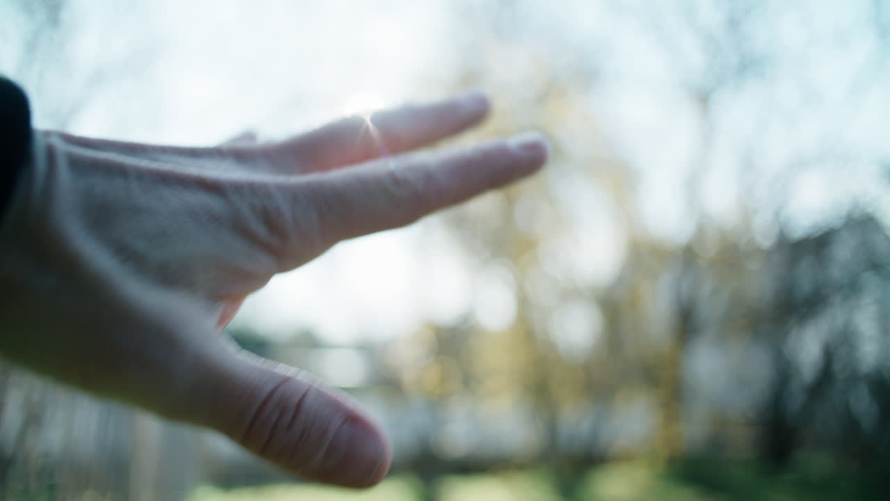 A point-of-view (POV) shot from a person's perspective looking at their own left hand. The hand is raised, holding a plain gold wedding ring towards the lens of a camera. The bright morning sun shines