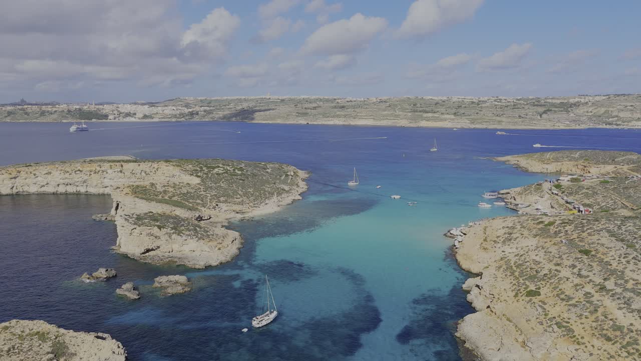 A high panoramic drone view over the Blue Lagoon shows Comino on the right, Gozo in the distance, calm turquoise water and several boats on a clear sunny day