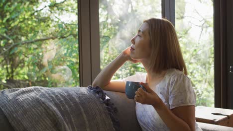 asian woman looking through window and lying on couch with mug of coffee