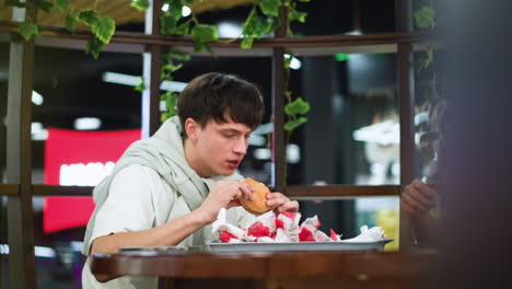 two colleagues interacting one eats burger on tray, camera capturing facial expressions and hand gestures, burger dripping sauce, second colleague listening attentively and smiling