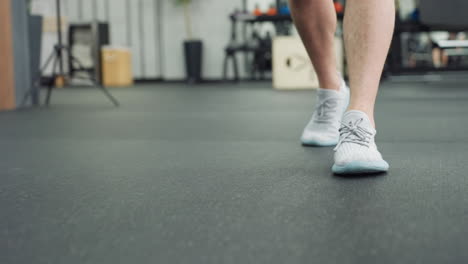 leg view of fitness trainer moving across rubber floor past blurred equipment, light gray sneakers stepping with purpose, highlighting active lifestyle motivation in modern training environment