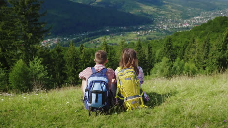 Young-Couple-Of-Tourists-With-Backpacks-Sitting-On-A-Green-Meadow-Looking-At-A-Picturesque-Montaña