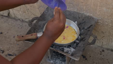 African-woman-cooks,-prepares-an-egg-omelette-in-a-frying-pan-on-a-charcoal-stove,-to-accompany-with-banku,-a-ghanaian-dish